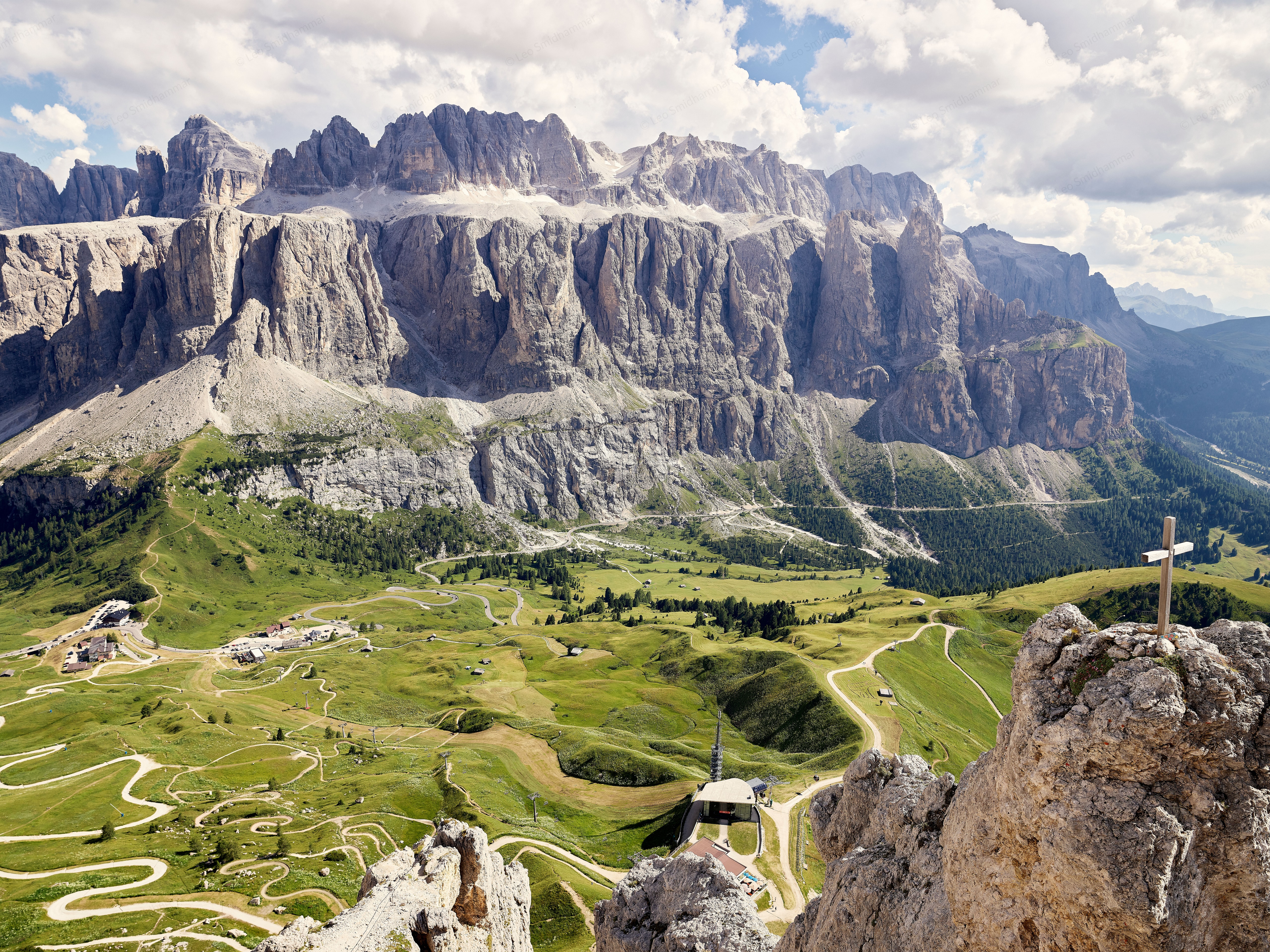 A panoramic view from the Cir peaks across green Alpine meadows and winding trails toward the immense rocky walls of the Sella massif, beneath a sky filled with summer clouds.