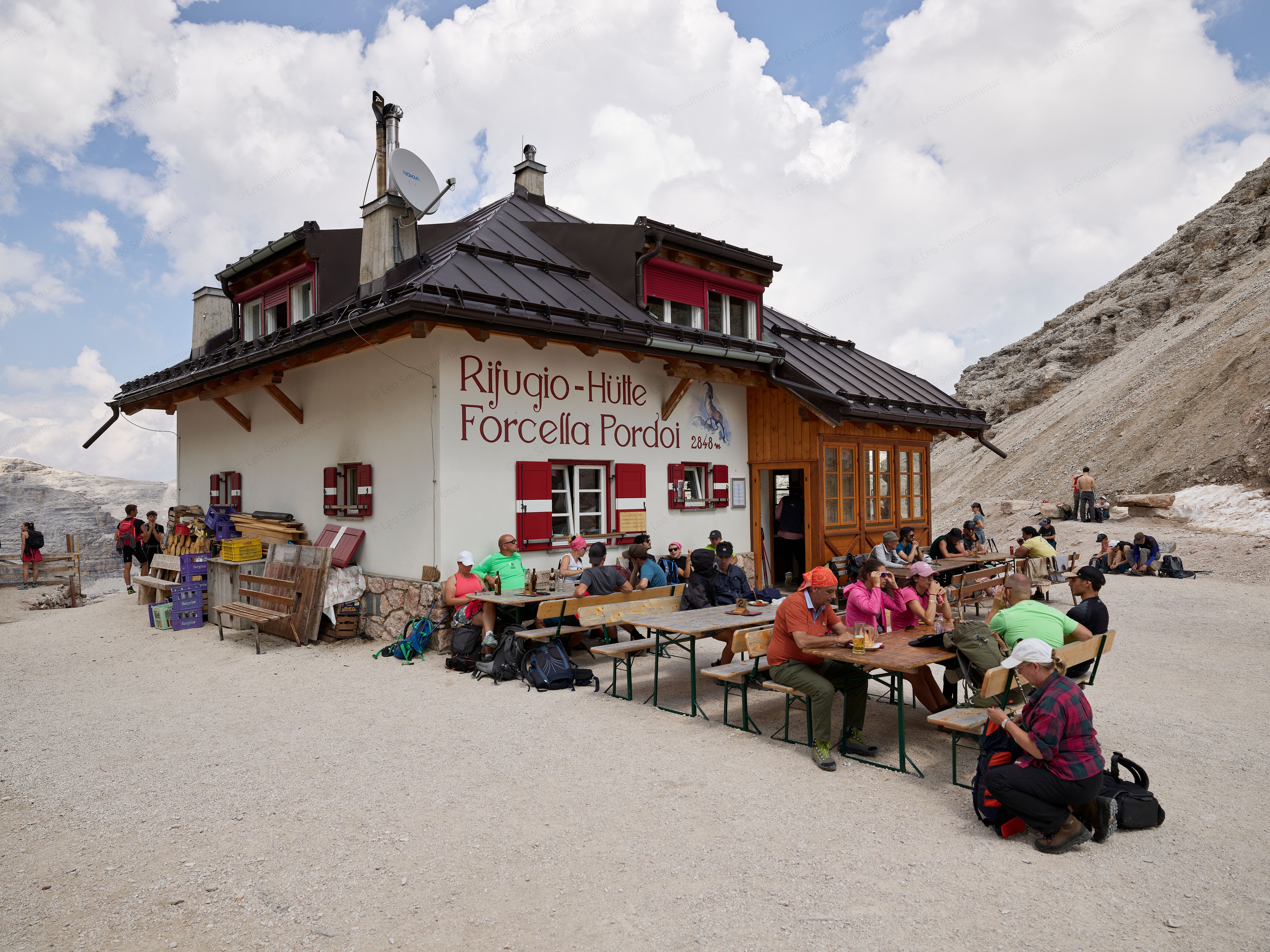 Hikers gather at outdoor tables beside Rifugio Forcella Pordoi, surrounded by pale rocky slopes beneath towering summer clouds.