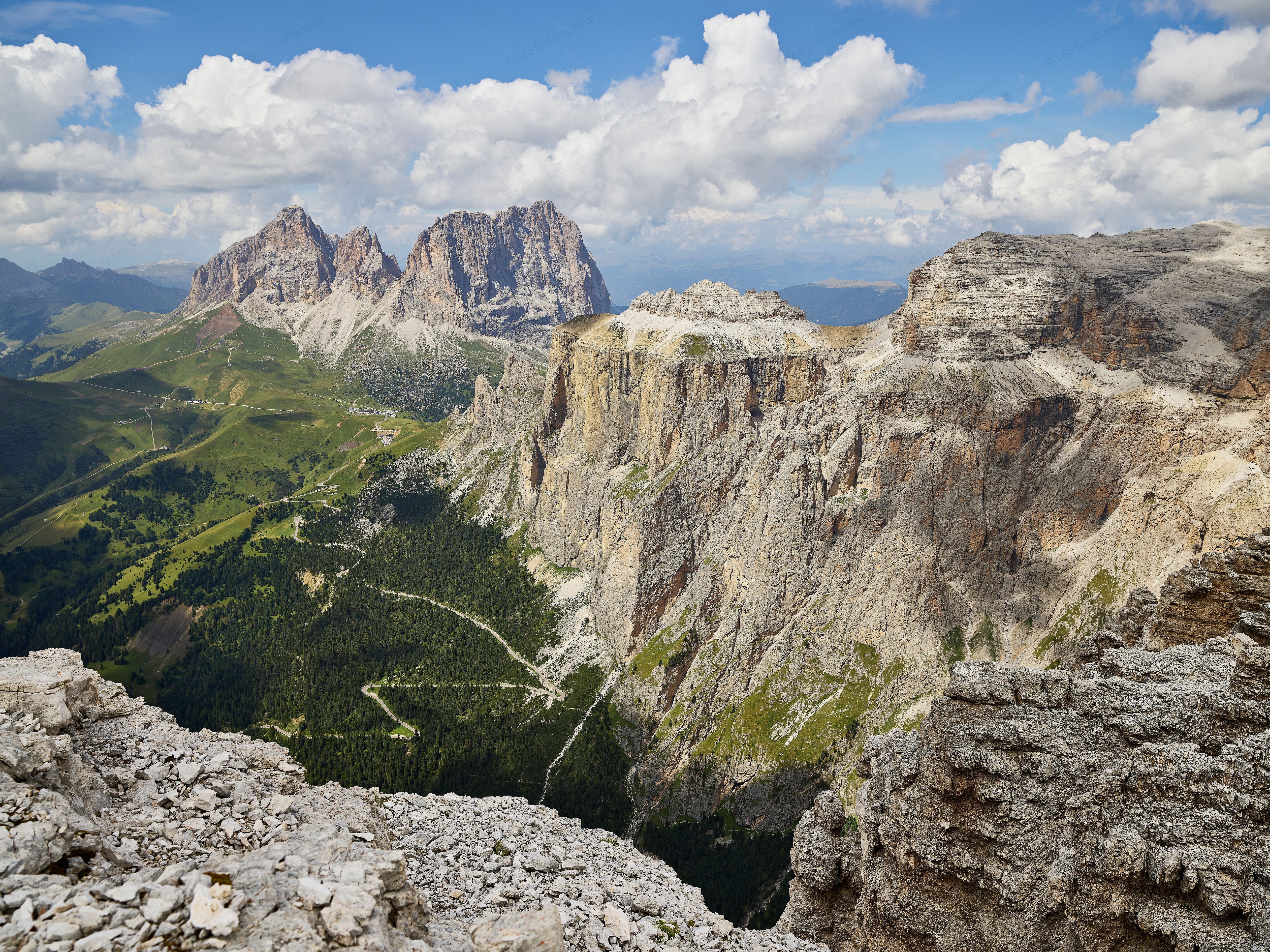 A panoramic view from Passo Pordoi across sheer limestone cliffs, forested valleys, and winding mountain roads toward the distant Sassolungo peaks beneath towering summer clouds.