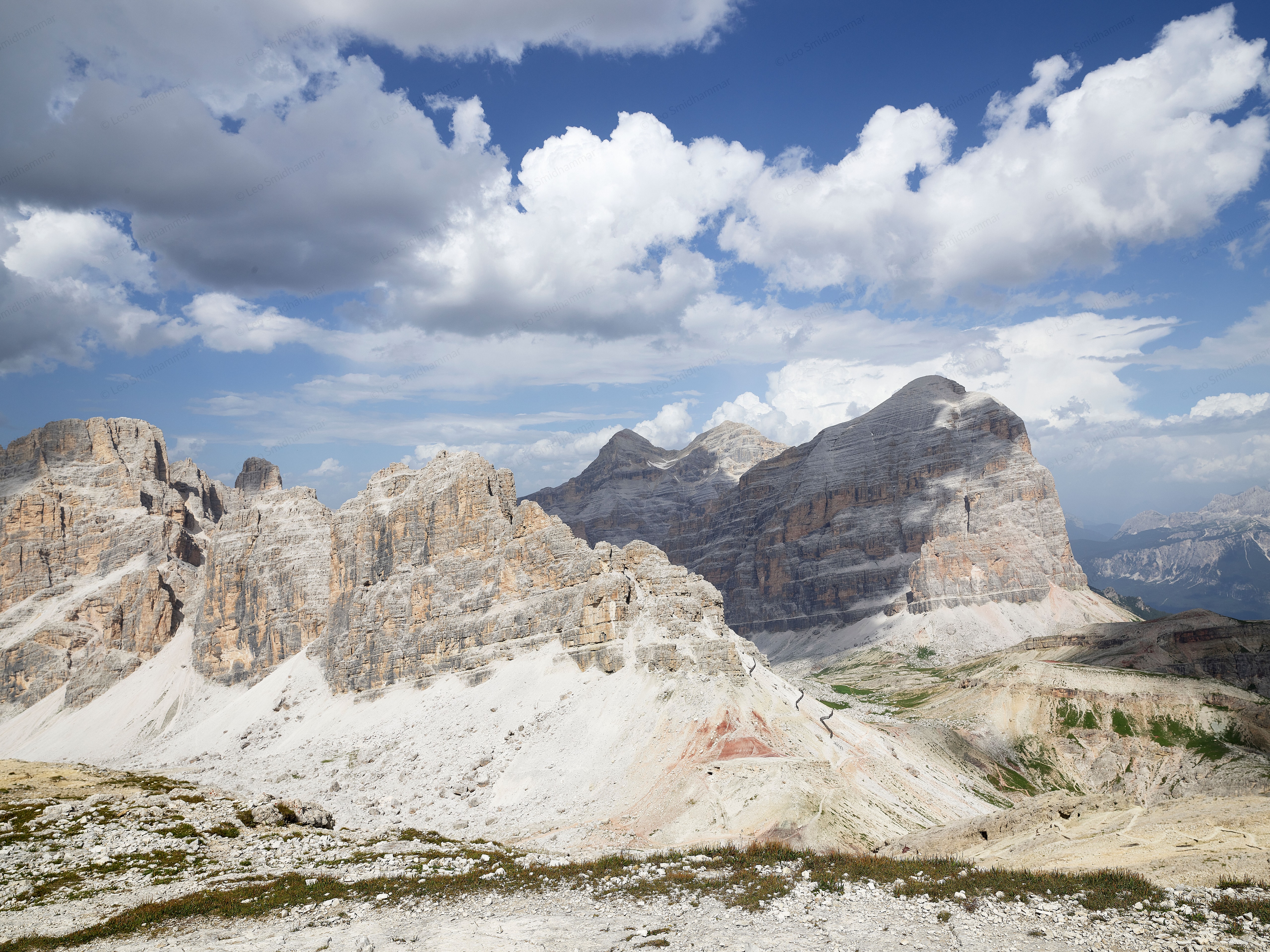 Pale Dolomite stone peaks and rugged ridges rise above a rocky Alpine landscape between Lagazuoi and Fanes, beneath dramatic white clouds and a deep blue summer sky.